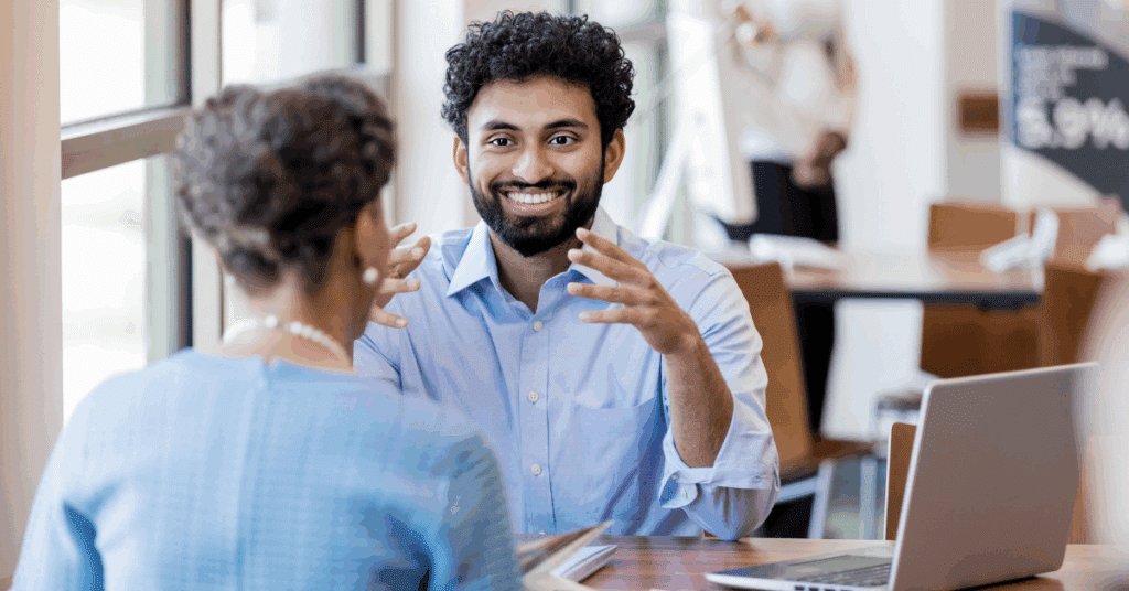 Cheerful young male bank employee gestures as he explains the bank's services with a mature female customer.