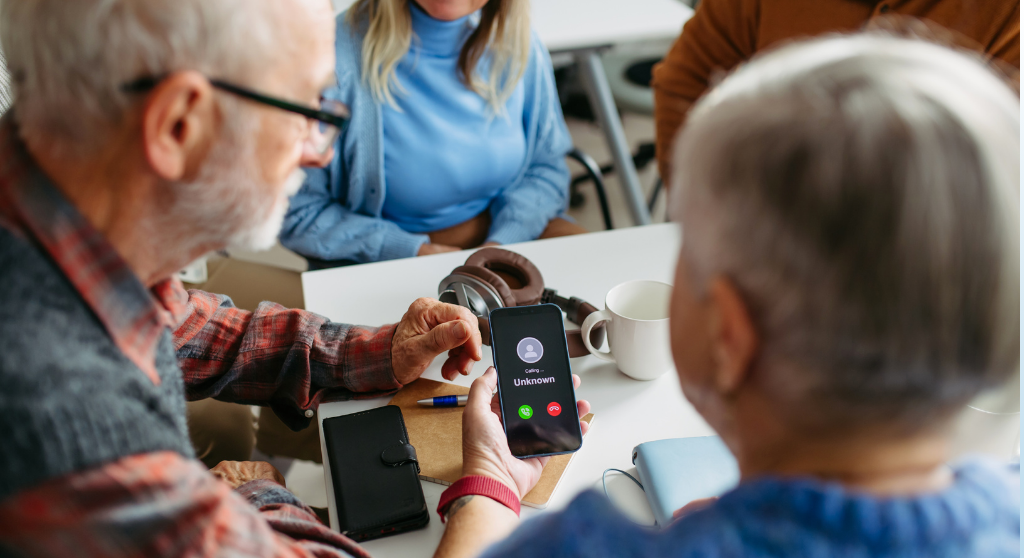 Older adults sit around a table as one person holds a smartphone displaying an incoming call labeled as "Unknown".