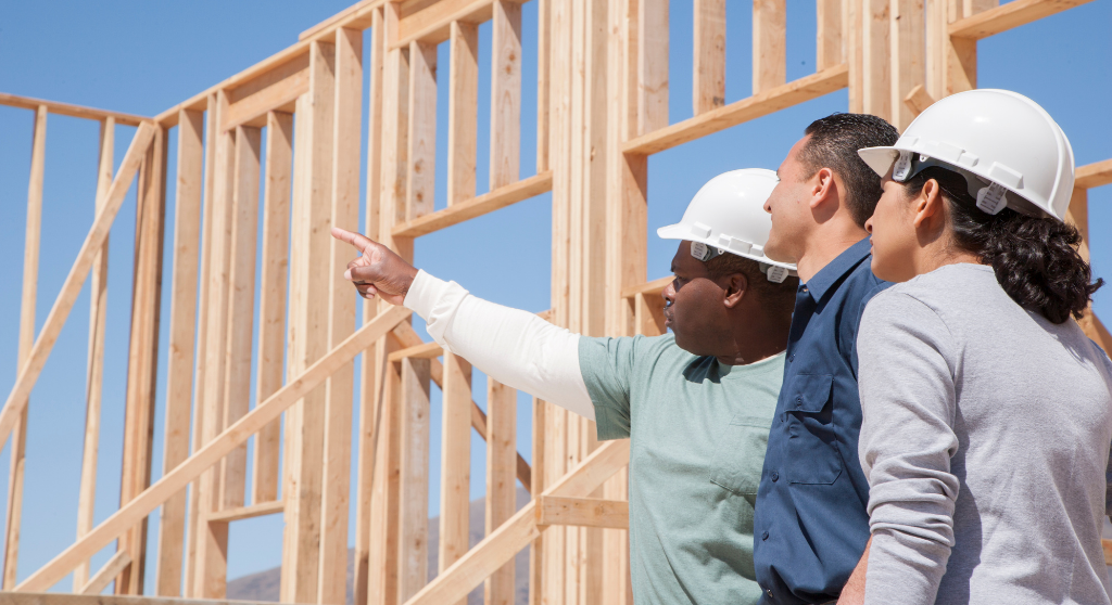 Three construction workers wearing hard hats stand in front of a wooden building frame, with one person pointing towards the frame.