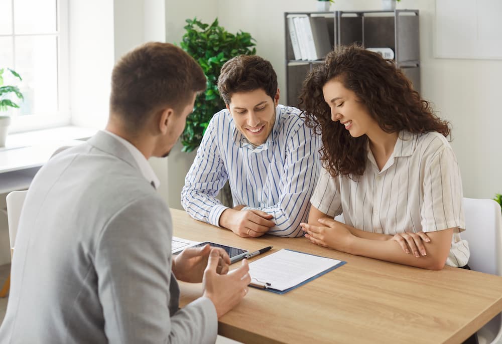 A banker sits across a desk from a smiling couple, reviewing paperwork together.